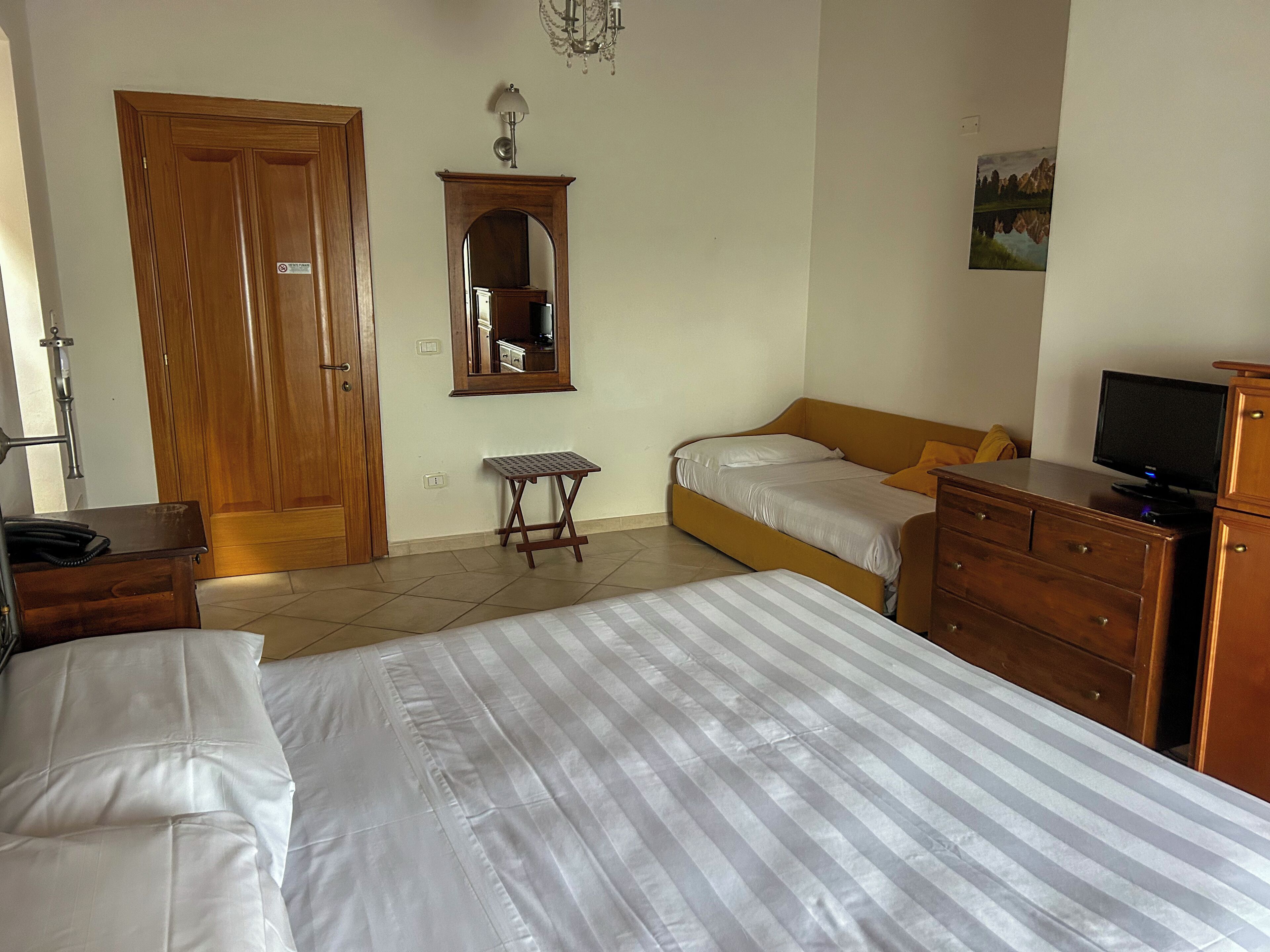 A bedroom with two beds, a wooden wardrobe, a small table, a mirror, a television, and a painting on the wall.
