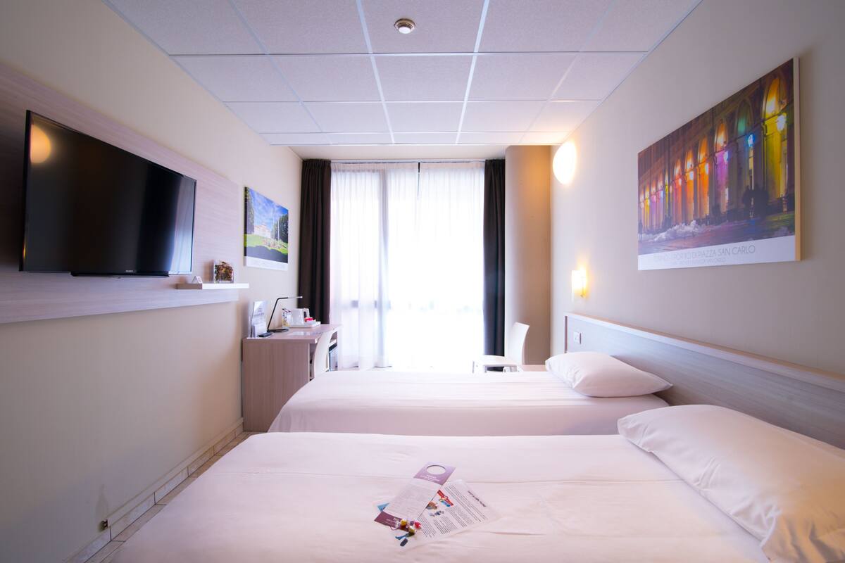 A hotel room with two beds, a flat-screen TV mounted on the wall, a desk with a chair, and a large window with curtains.