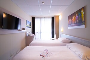 Habitación de hotel con dos camas, un televisor de pantalla plana montado en la pared, un escritorio con una silla y un ventanal con cortinas.