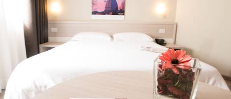 A hotel room with a neatly made bed, a bedside table with a pen and a card, a glass vase with flowers, and a wall-mounted picture of a building.