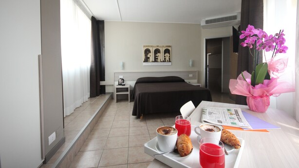 Ein Hotelzimmer mit einem Bett, einem Nachttisch mit Lampe, einem Tablett mit Frühstücksangeboten und einem Fenster mit Vorhängen.