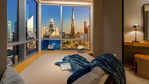 Une chambre avec une grande fenêtre donnant sur le paysage urbain, un lit avec une couverture, une table de chevet avec une lampe et un petit vase avec des fleurs.