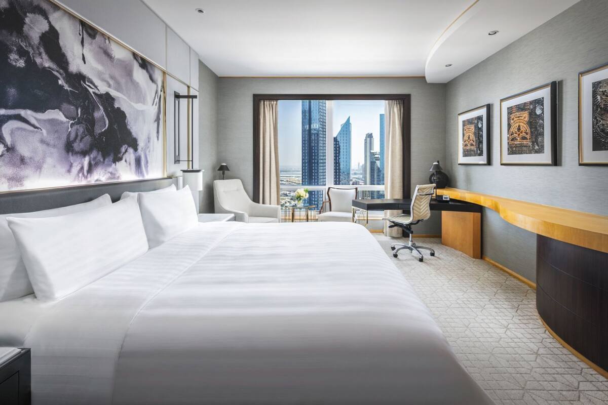 A modern hotel room with a large bed, a desk, a chair, and a view of tall buildings.