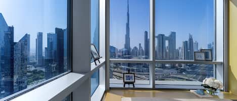 A high-rise room with a large window offering a cityscape view, a small table with a book and a vase of flowers, and a framed picture on the windowsill.