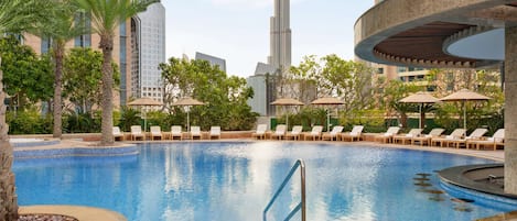 Una piscina con sillones de descanso y sombrillas, rodeada de palmeras y edificios modernos.