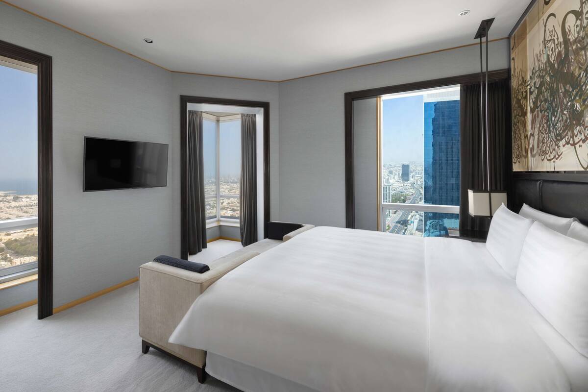 A modern hotel room with a large bed, a flat-screen TV, a seating area with a view of the city, and a large window offering a panoramic view.