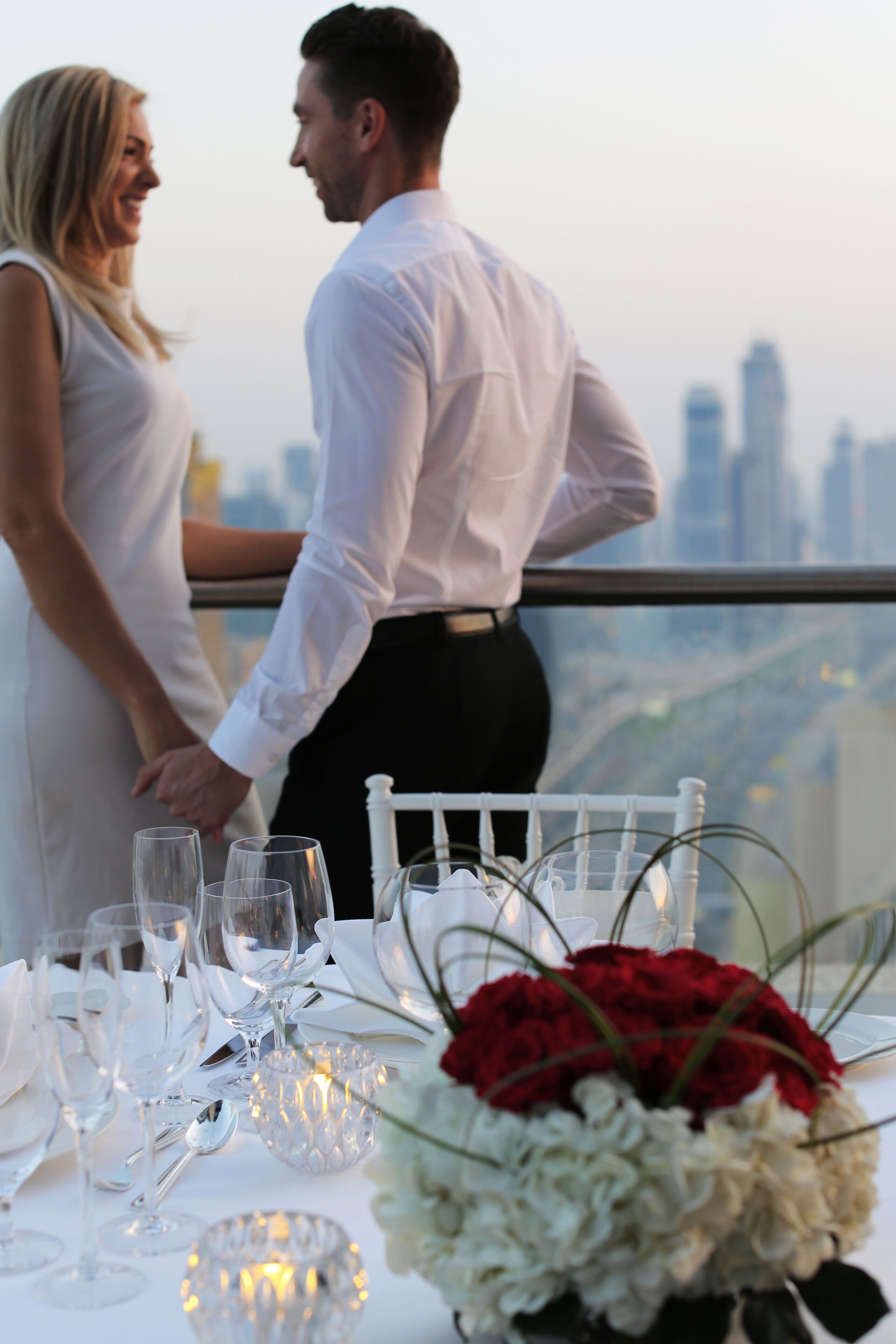 Una pareja de pie junto a una mesa, tomados de la mano, con vistas a la ciudad.