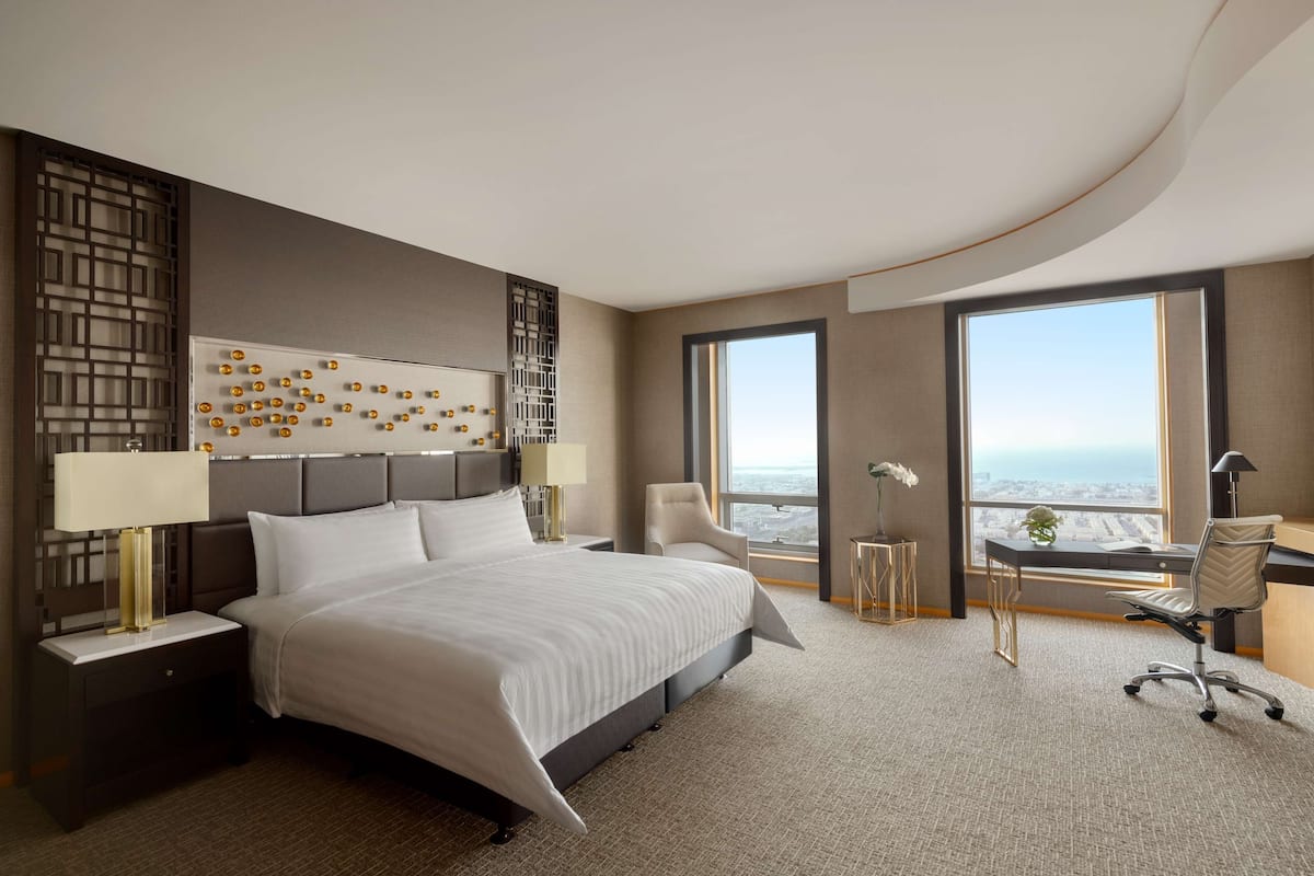 A modern hotel room with a large bed, two bedside tables with lamps, a desk with a chair, and large windows offering a city view.