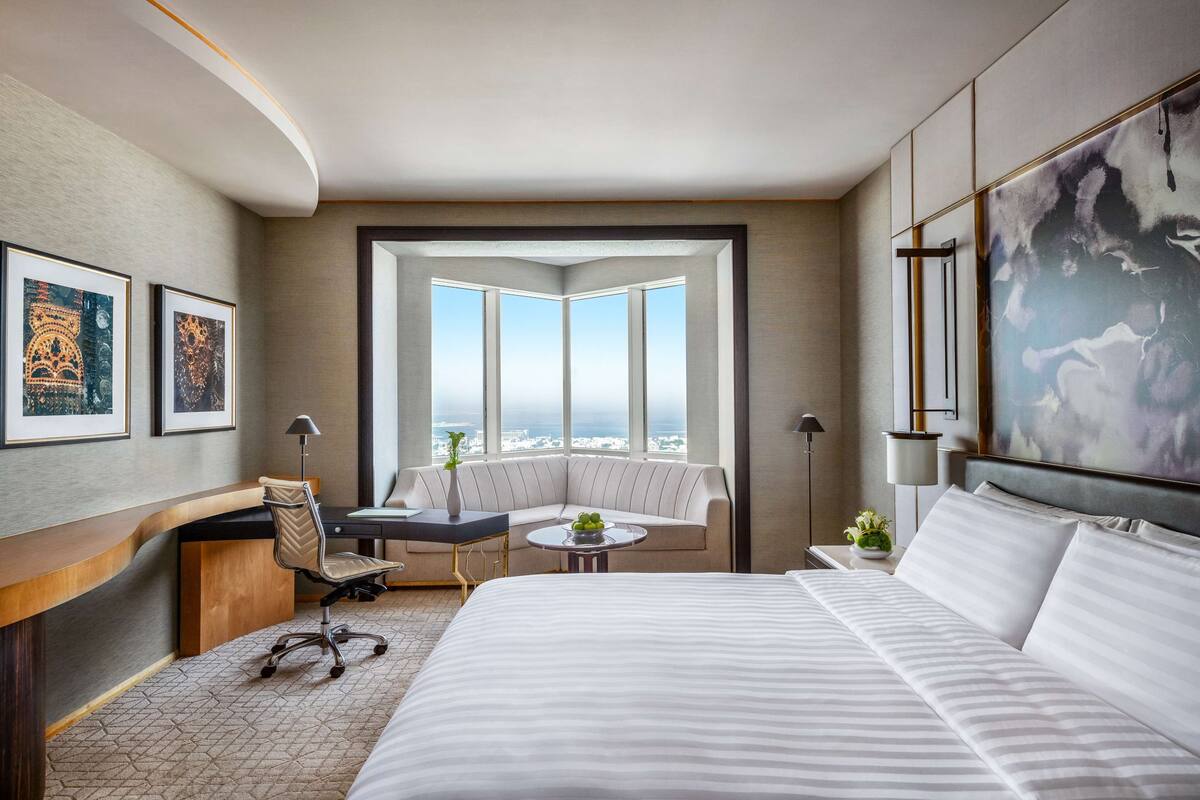 A hotel room with a large bed, a desk, a chair, a small table, and a sofa. There is a large window with a view of the city.
