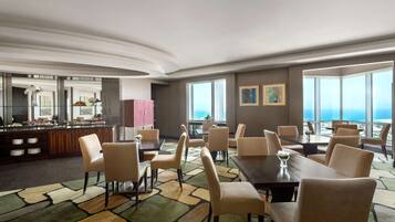 A hotel dining area with multiple tables and chairs, a buffet counter, and large windows offering a view of the city.