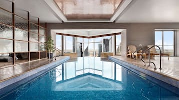 Une piscine intérieure moderne avec des parois en verre transparent, une terrasse en bois et une vue sur les bâtiments de la ville.