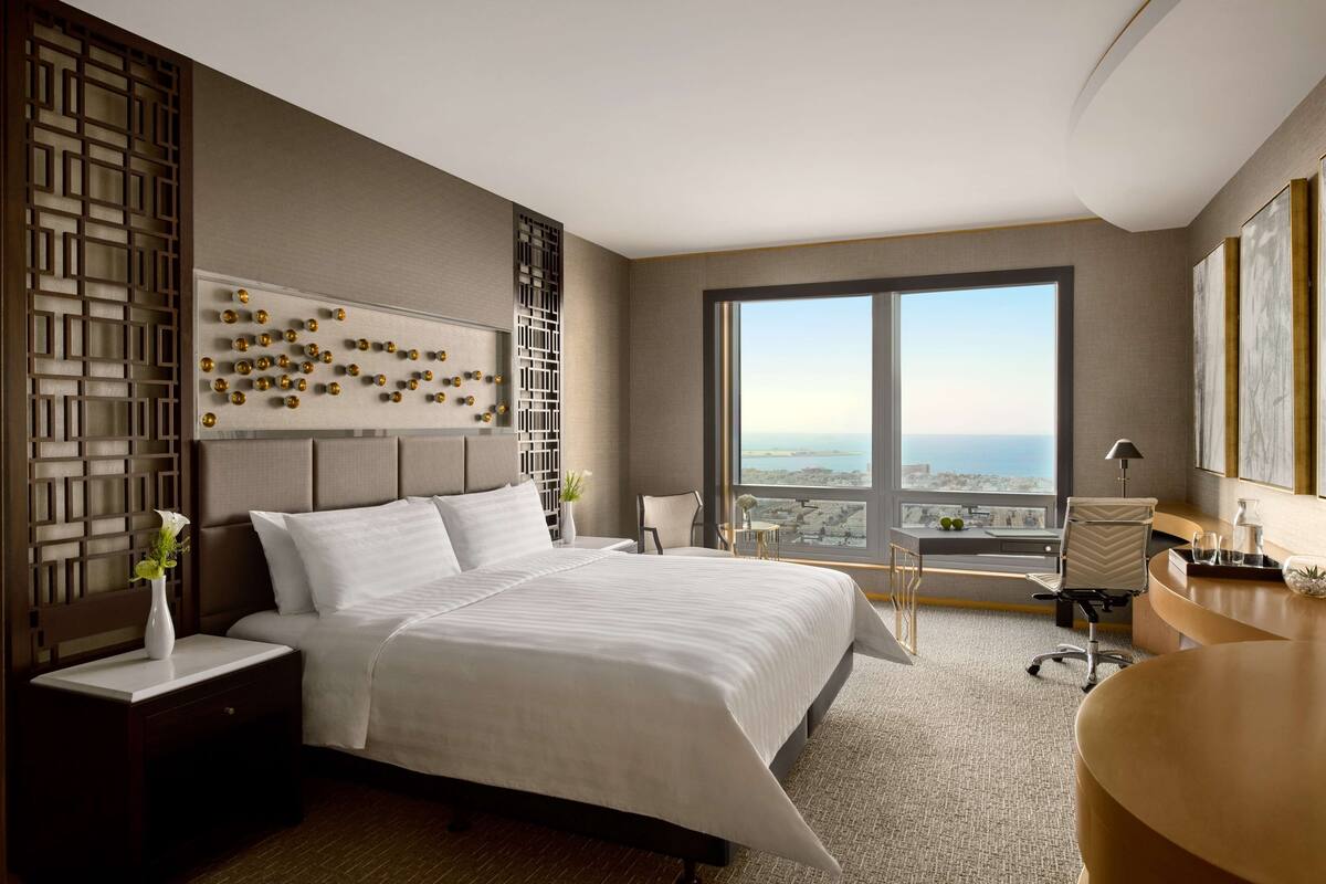 A modern hotel room with a large bed, a desk, a chair, and a view of the city.