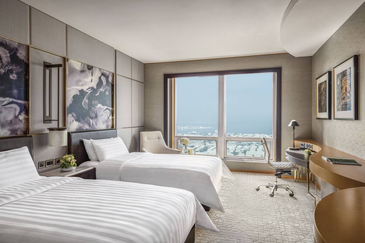 A hotel room with two beds, a desk, a chair, and a large window offering a city view.