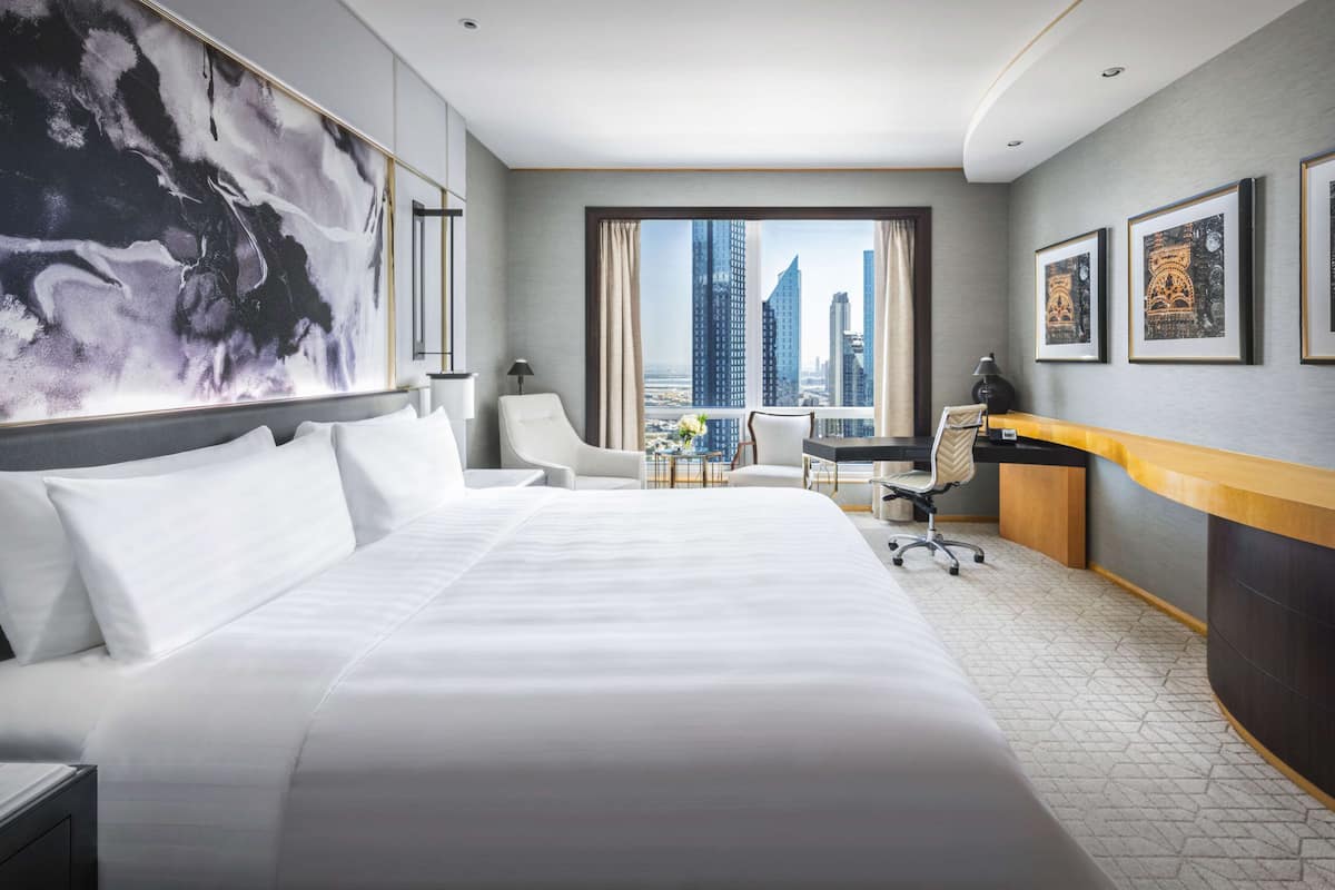 A modern hotel room with a large bed, a desk, a chair, and a view of tall buildings.