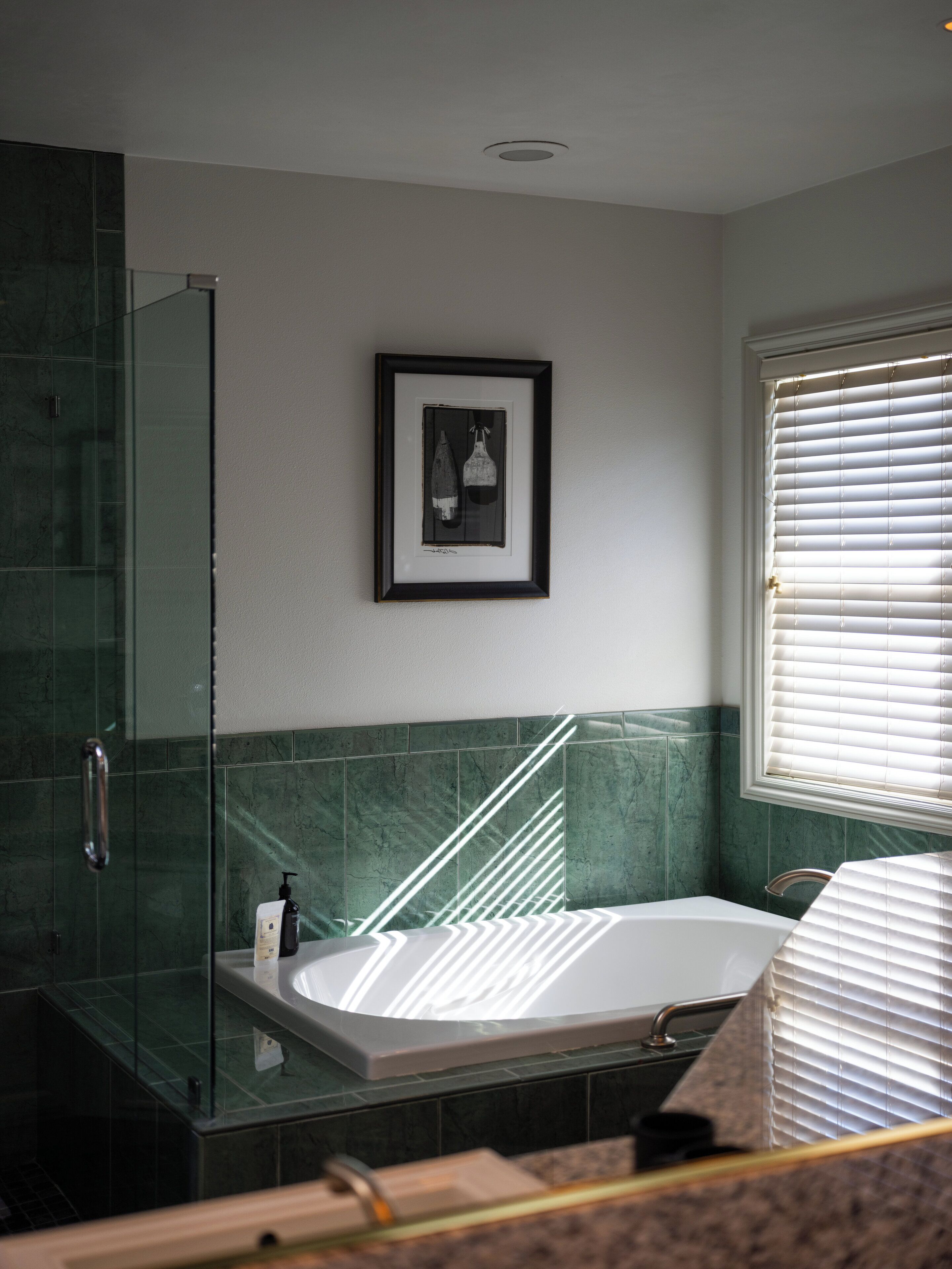 A modern bathroom with a glass shower enclosure, a framed artwork above the bathtub, and a window with blinds.