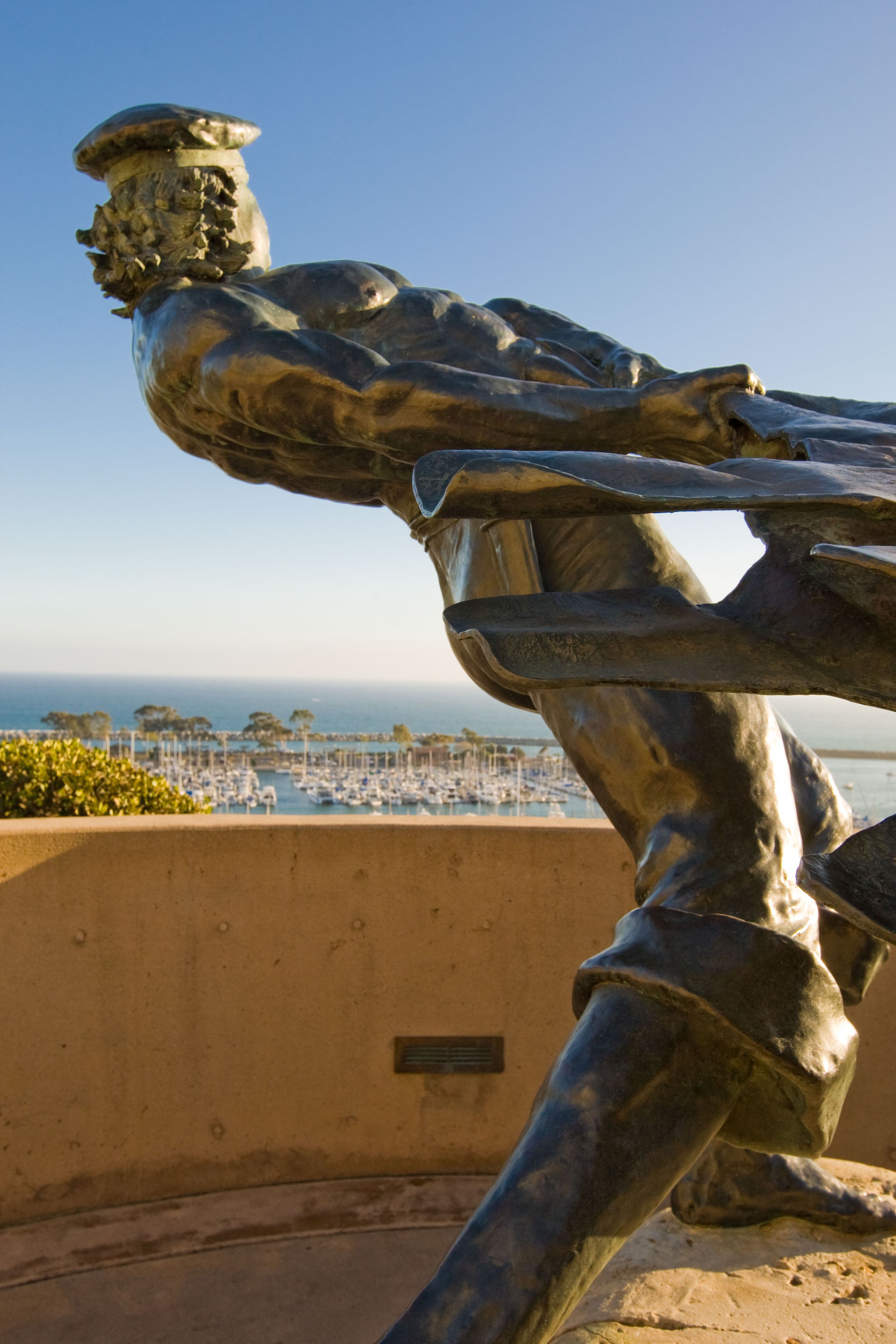 A bronze statue of a muscular figure with a raised arm, overlooking a marina with boats and a clear sky.
