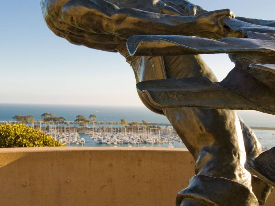 A bronze statue of a muscular figure with a raised arm, overlooking a marina with boats and a clear sky.