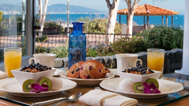 A breakfast table with plates of food, a blue vase, and a view of the sea. - Blue Lantern Inn, A Four Sisters Inn (Dana Point)