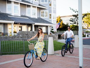 Two people riding bicycles on a paved path with a stone wall and a building in the background.