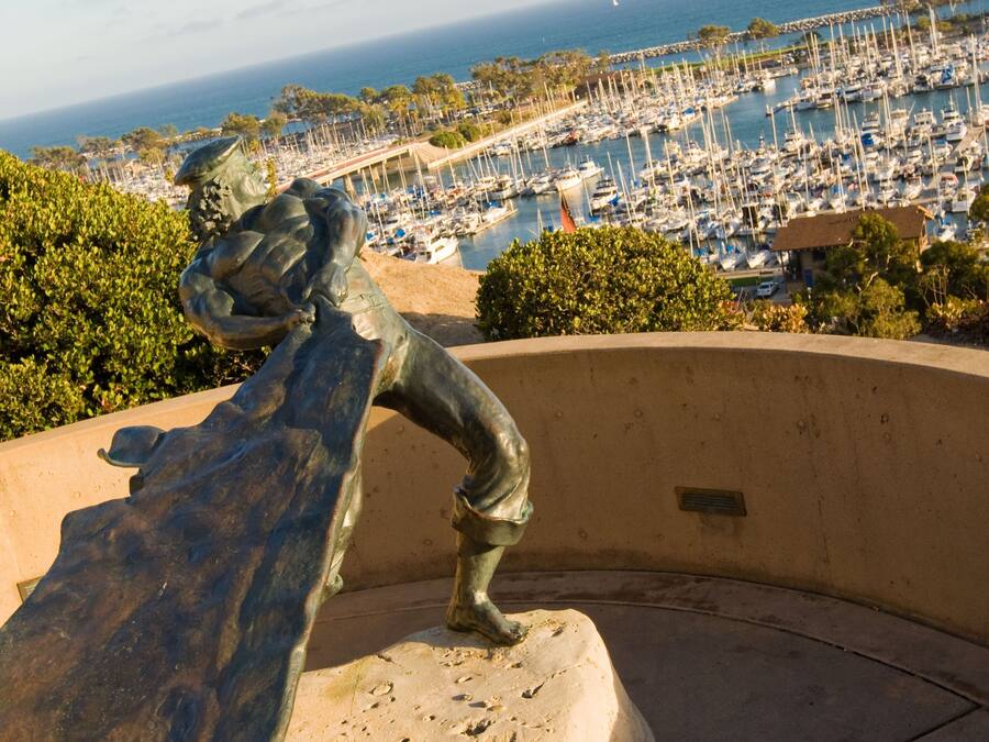 A sculpture of a figure on a curved bench overlooking a marina with boats and a clear view of the ocean.