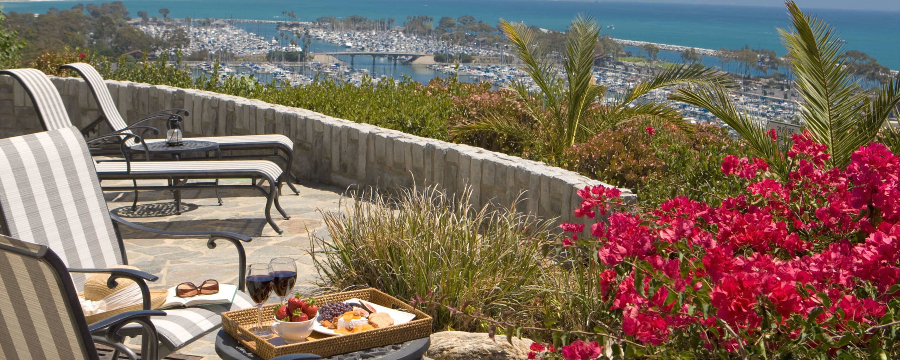 Une terrasse avec deux fauteuils de jardin, un plateau en osier contenant des collations et des boissons, et une vue sur une marina et l’océan.