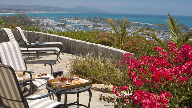 A terrace with outdoor seating, a table with food and drinks, and a view of a coastal area with boats and a bridge.