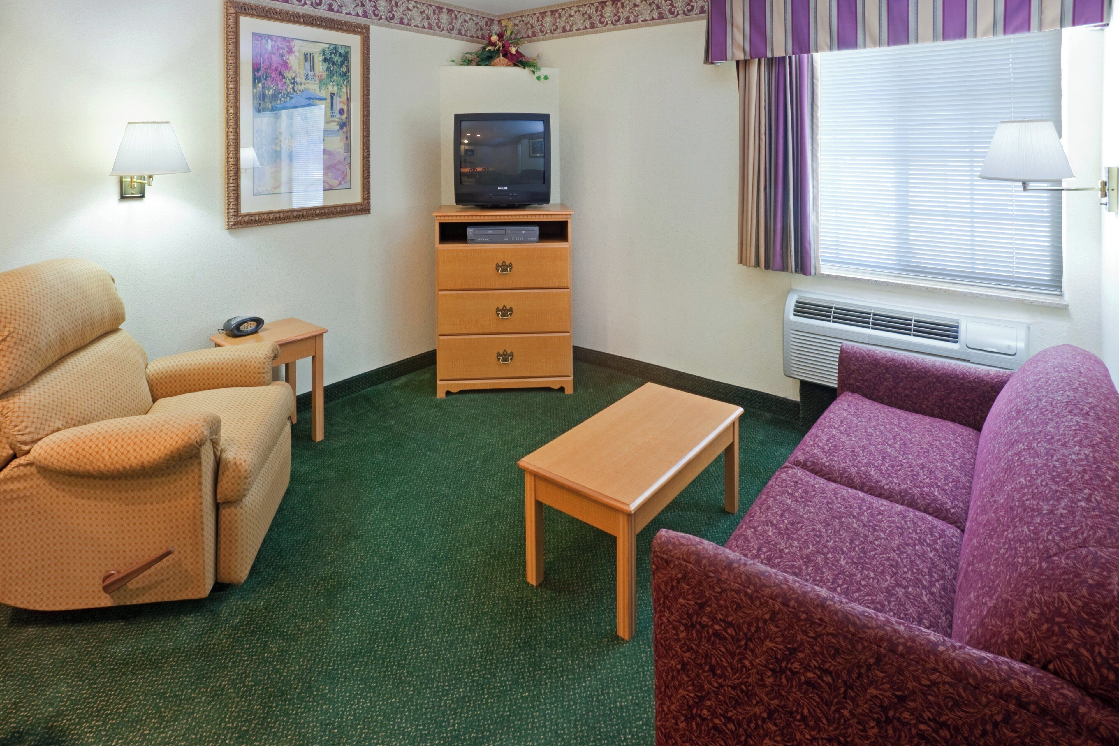 Une chambre meublée comprenant un canapé, un fauteuil, une petite table, un téléviseur posé sur un meuble en bois et une fenêtre avec des stores.