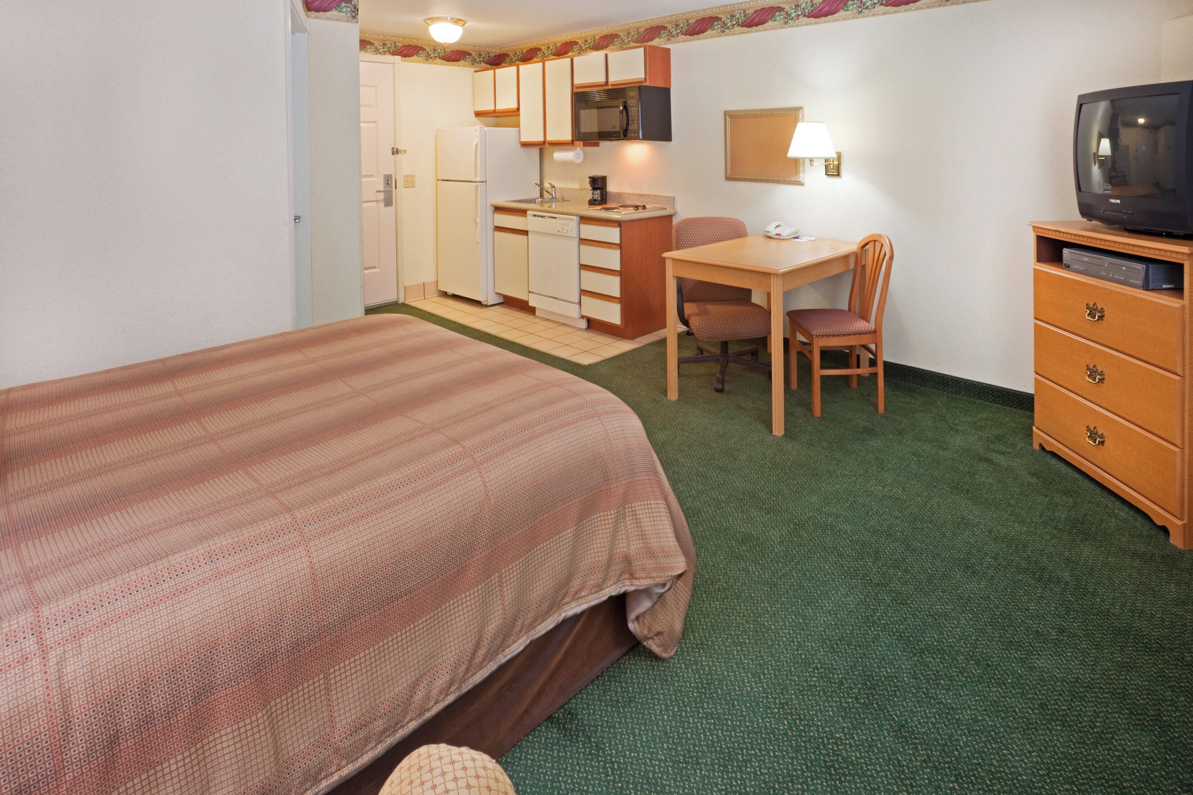 Een slaapkamer met een bed, een bureau, een stoel, een televisie en een kitchenette.