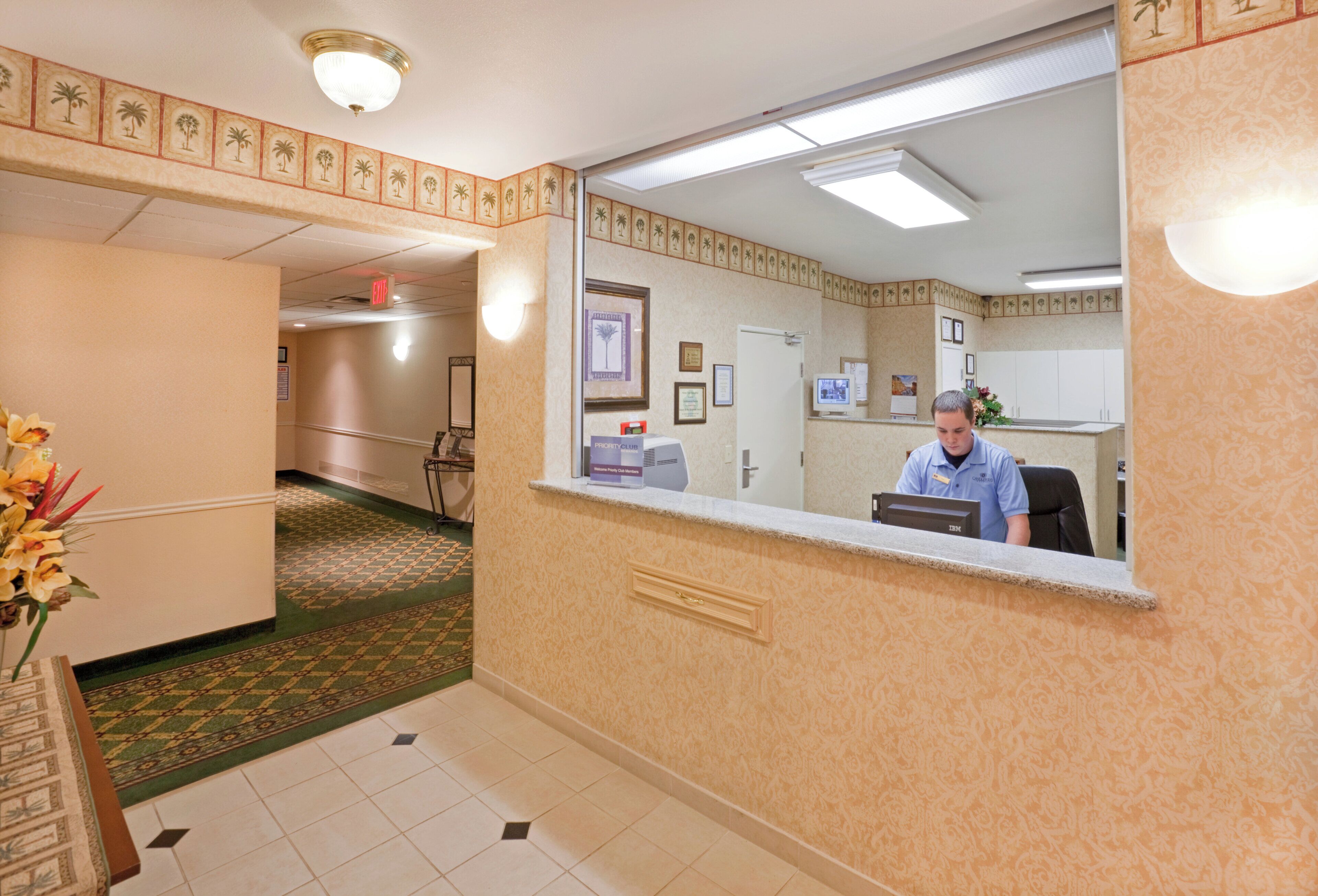 Un espace d’accueil d’hôtel avec un réceptionniste au comptoir, un bureau d’accueil, un ordinateur fixé au mur, des certificats encadrés et un tapis à motifs.