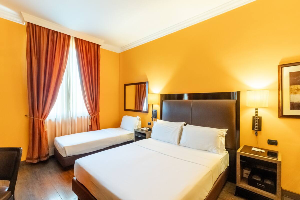 Una habitación de hotel con dos camas, un escritorio, una silla, una mesita de noche, una lámpara y un espejo.