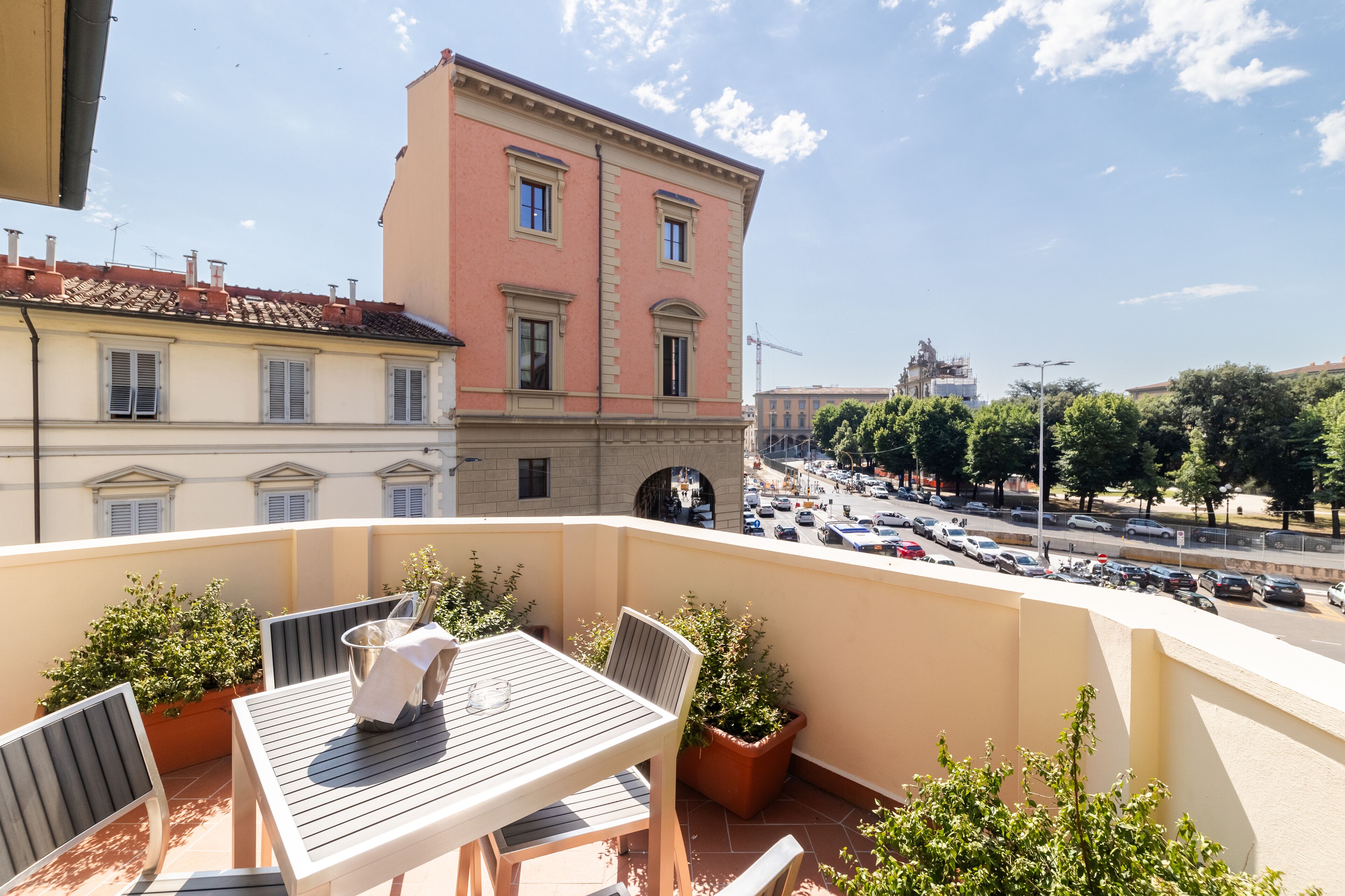 A balcony with a table and chairs, potted plants, and a view of a city street with buildings and parked cars.