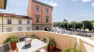 A balcony with a table and chairs, potted plants, and a view of a city street with buildings and parked cars.