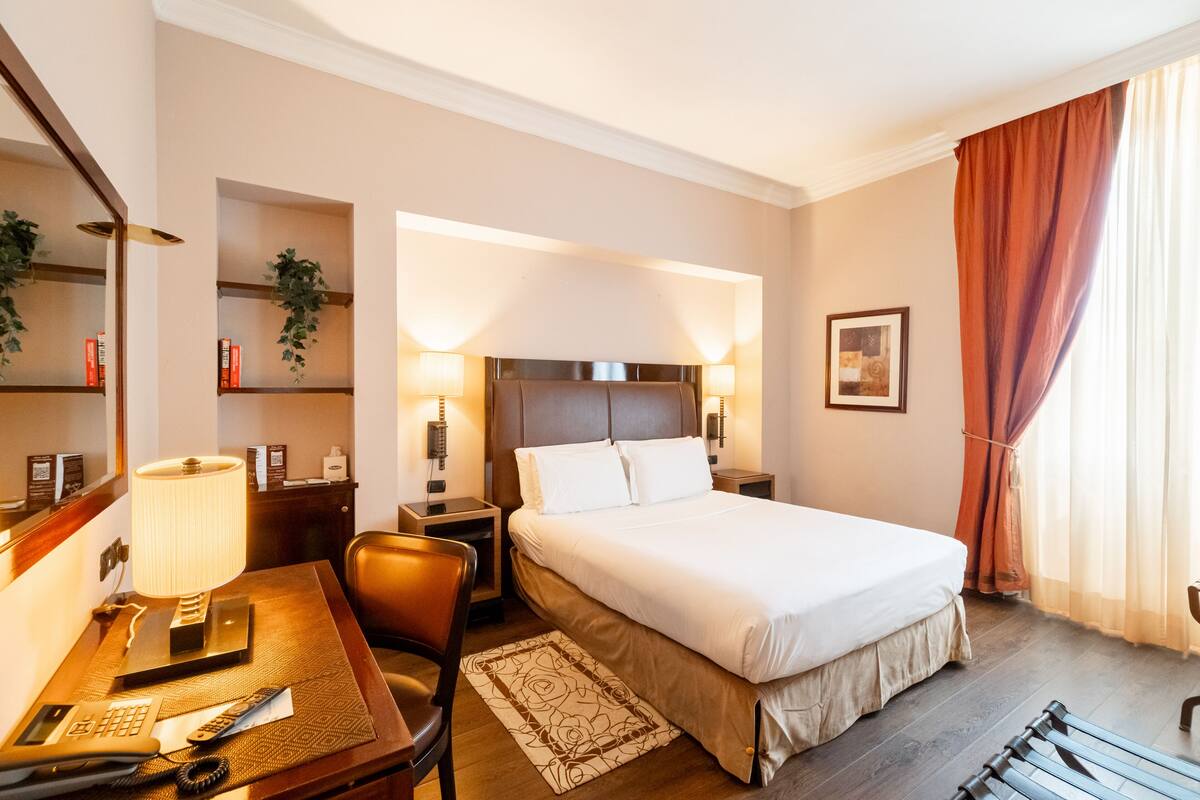 Habitación de hotel con una cama grande, lámparas de noche, un escritorio con teléfono, una silla y una lámpara de escritorio.
