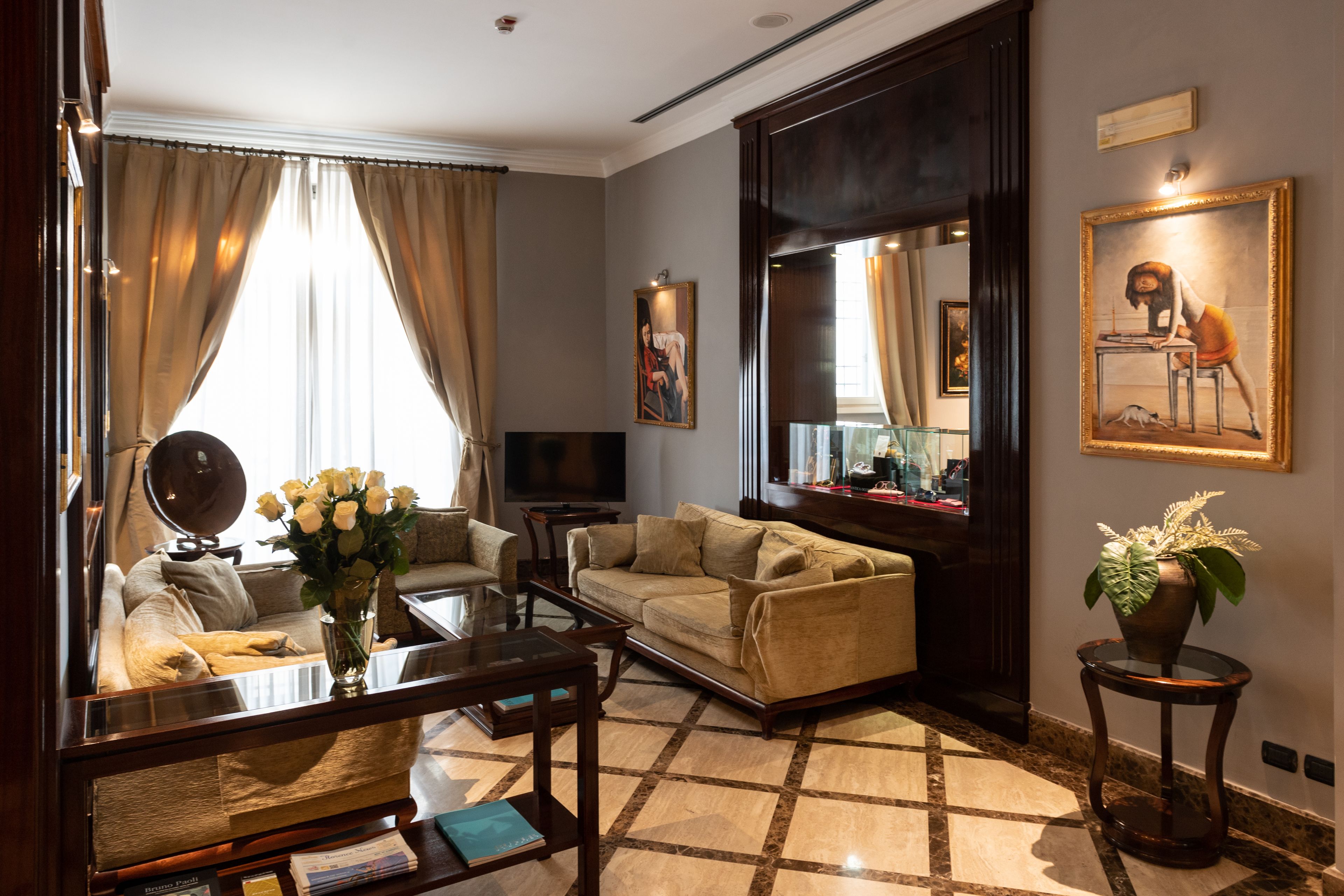 A spacious living room with a sofa, coffee table, and a TV. There are curtains, a vase with flowers, and a framed picture on the wall.