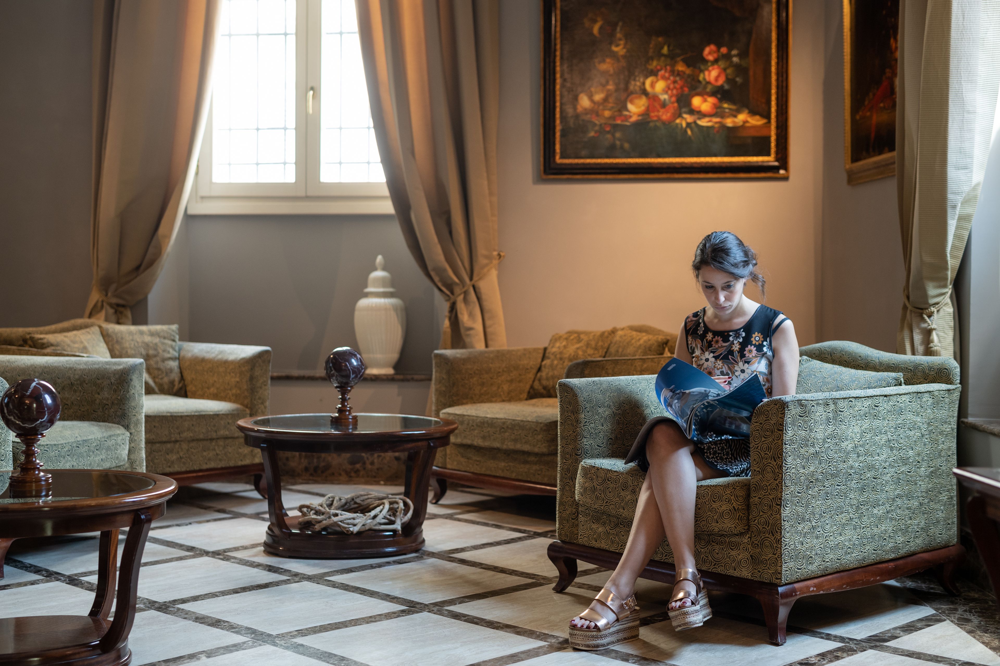 A woman sitting on a patterned armchair in a room with a checkered floor, a painting on the wall, and a window with curtains.