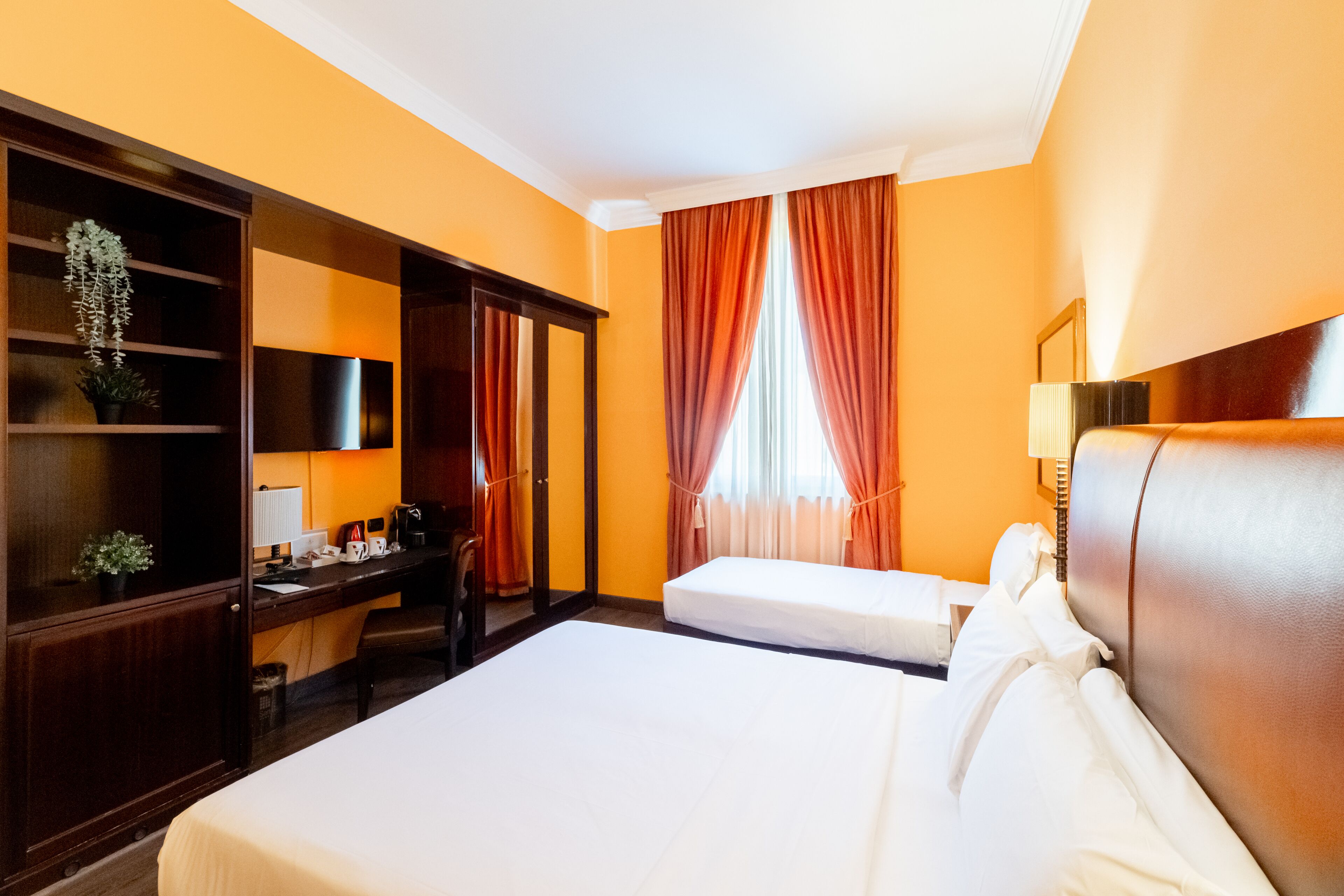 A hotel room with two beds, a desk, a chair, a wardrobe, and a window with curtains.