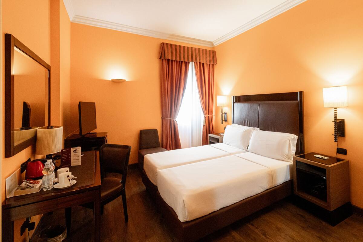 Habitación de hotel con dos camas, un escritorio con silla, un espejo, una mesita de noche con lámpara y una ventana con cortinas.