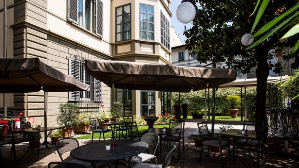 Un patio al aire libre con mesas y sillas, rodeado de plantas en macetas y un edificio con ventanas amplias.