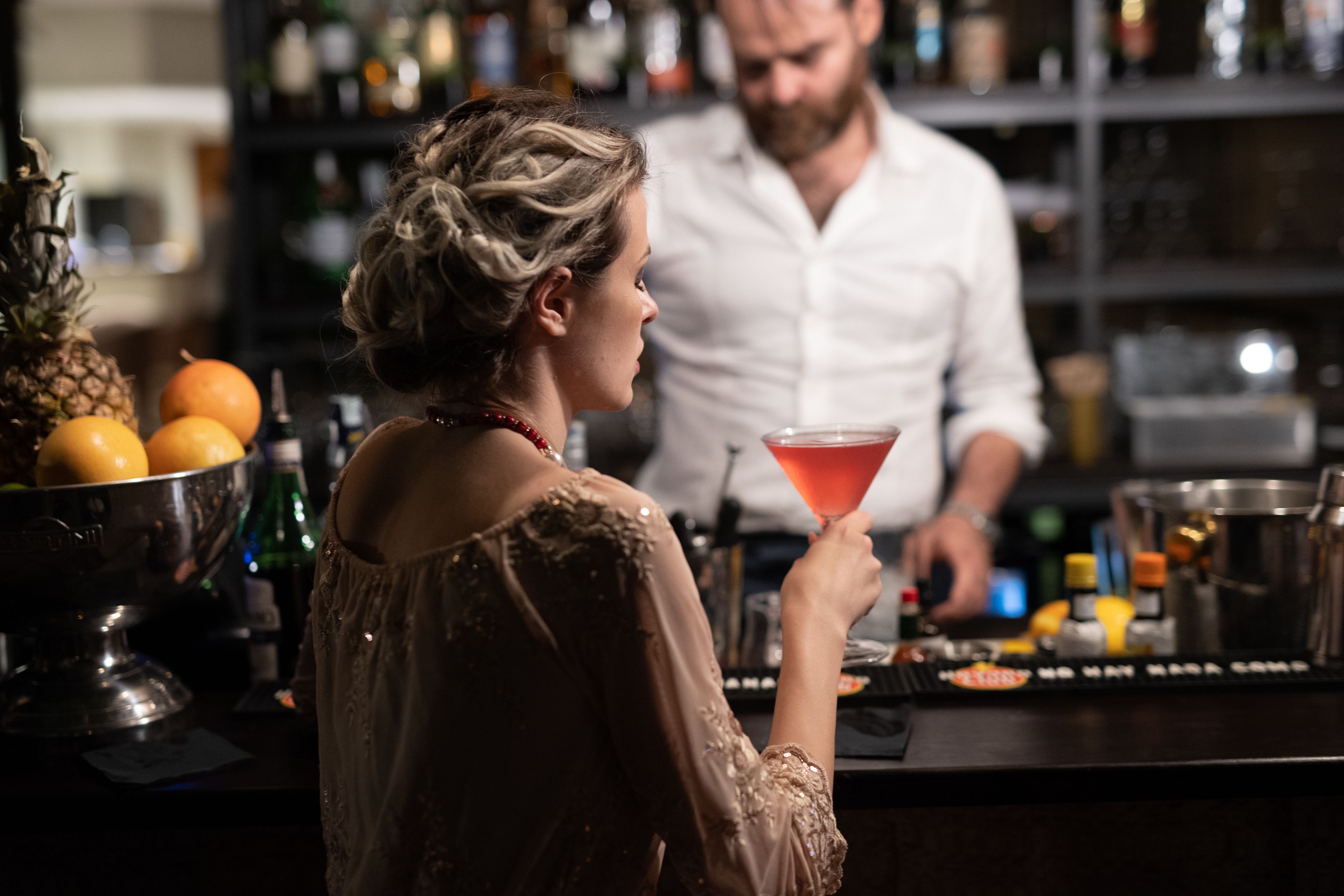 A bar area with a bartender preparing drinks. There are bottles and a fruit bowl visible.