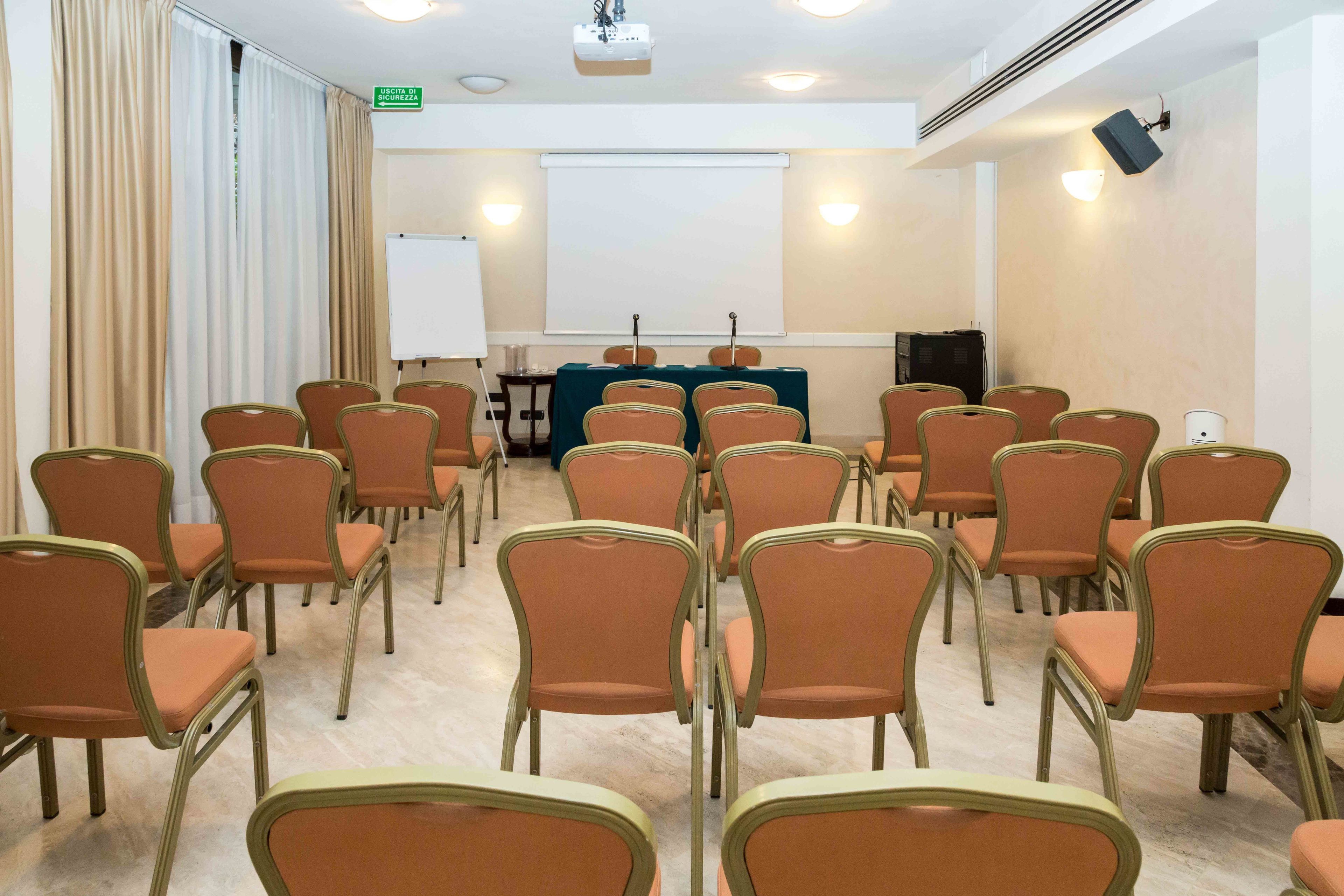 Une salle de conférence avec des rangées de chaises orange, un tableau blanc et un écran de projection.