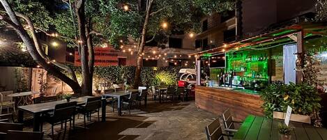 Un espace extérieur aménagé en terrasse avec des tables et des chaises, un bar dont l’intérieur est illuminé de lumière verte, et des guirlandes lumineuses.