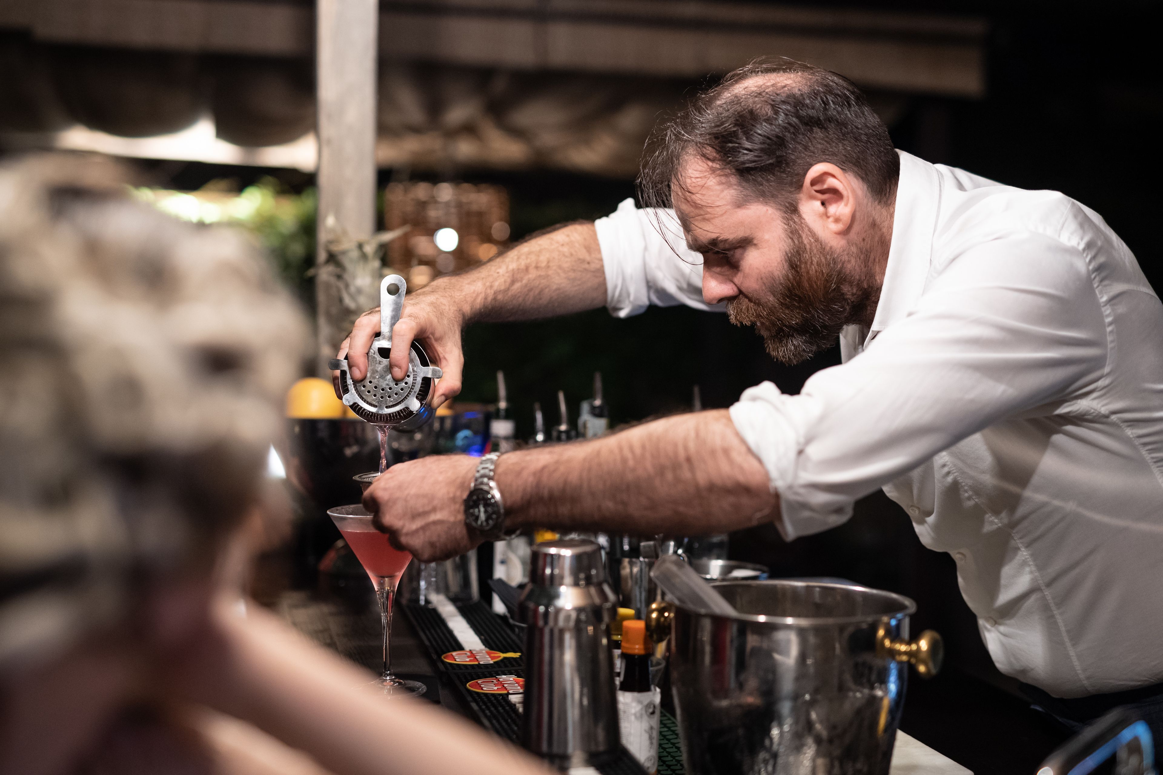 En bartender laver en drink bag baren, med forskellige flasker og en stor metalbeholder.