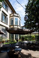 Patio esterno con tavoli e sedie rotonde, circondato da piante in vaso e un grande ombrellone.