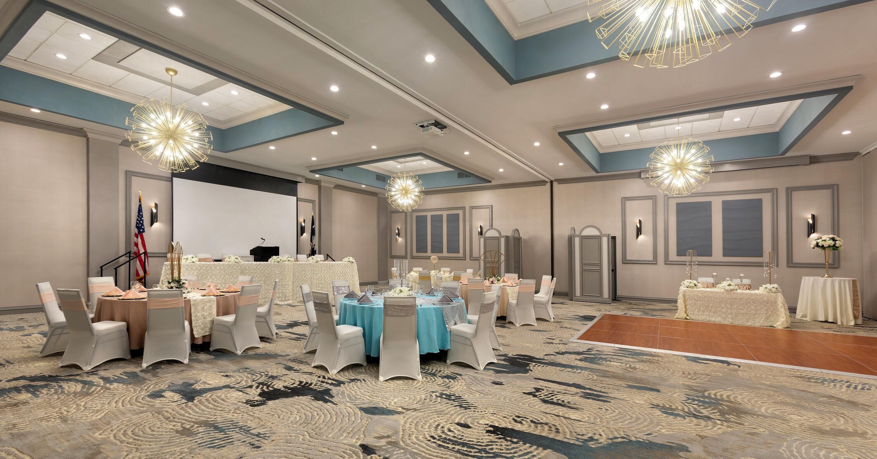 A spacious banquet hall with round tables, chairs, and a stage area.