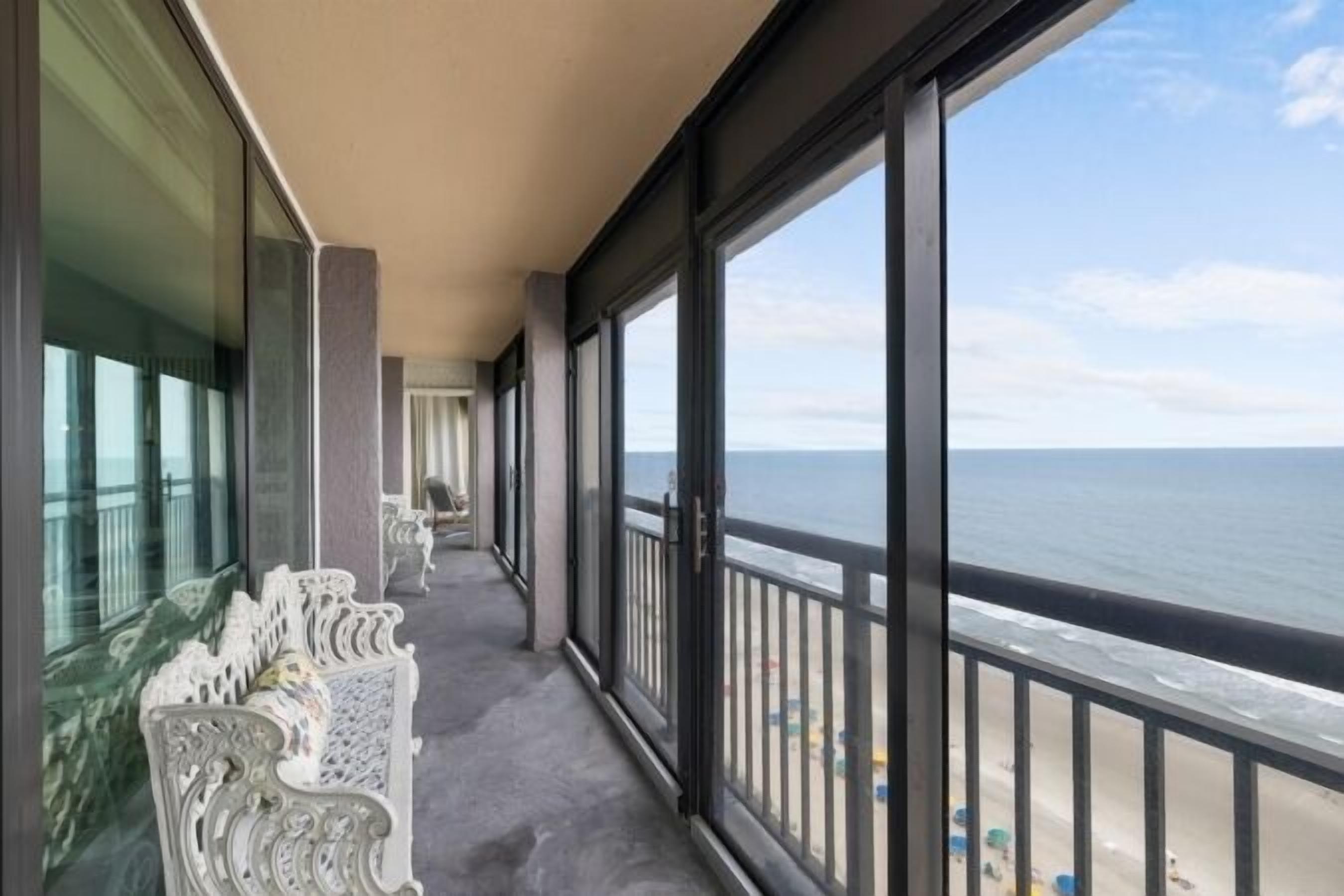 A balcony with a view of the ocean, white ornate chairs, and a glass railing.