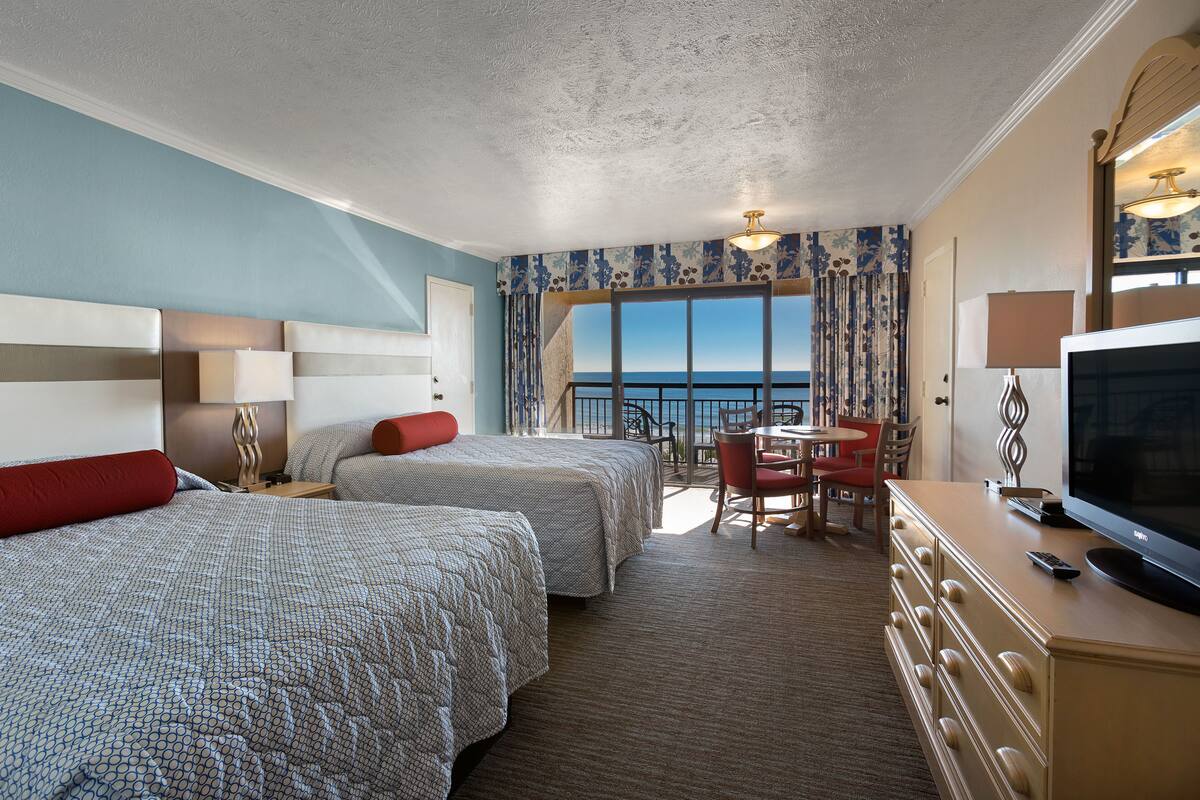 A hotel room with two beds, a TV, a desk, and a view of the sea.
