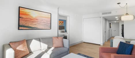 A modern living room with a sofa, a coffee table, and a chair. There is a painting on the wall and a door leading to another room.