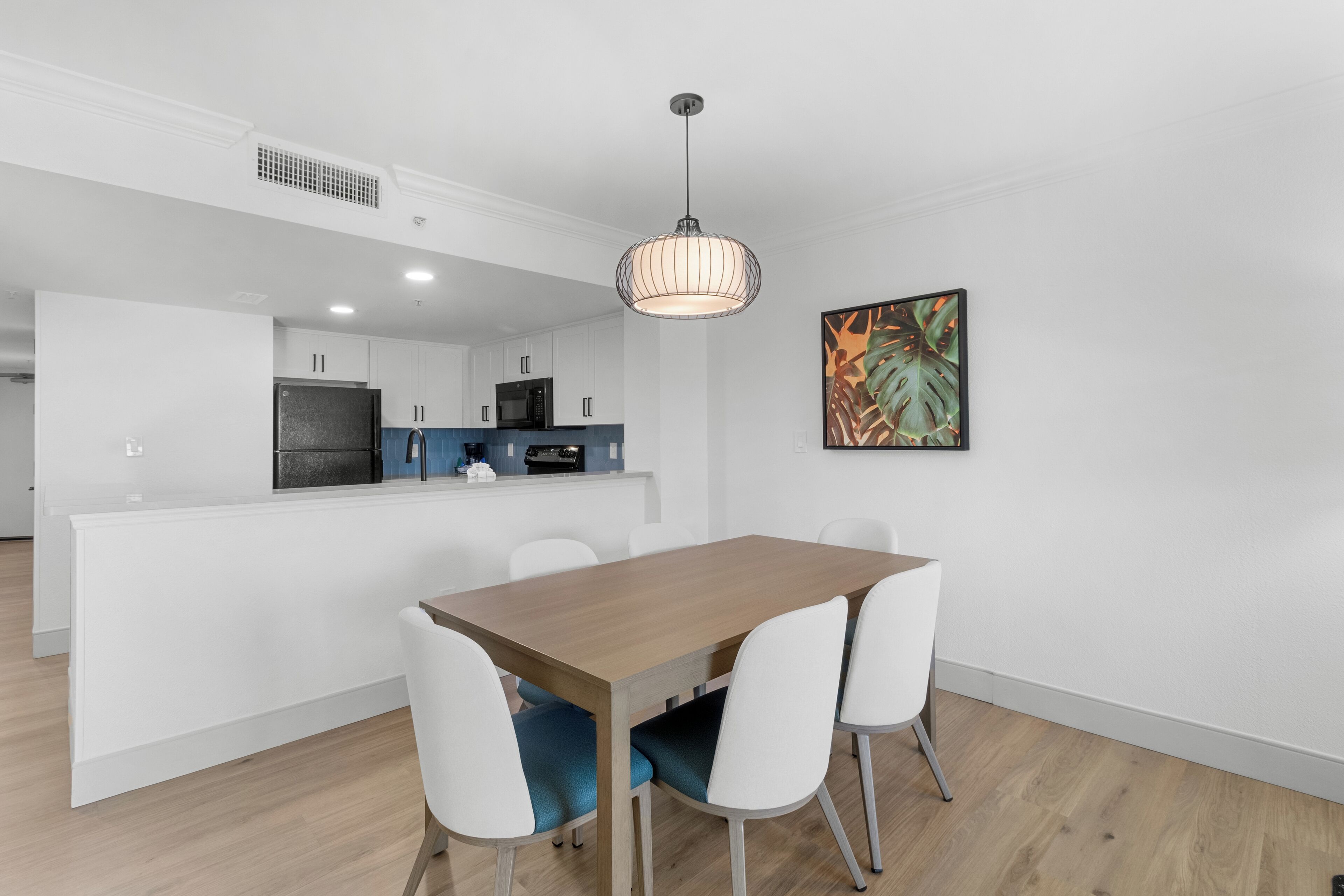 A modern kitchen with white cabinets, a black refrigerator, and a microwave. There is a dining area with a wooden table and white chairs.