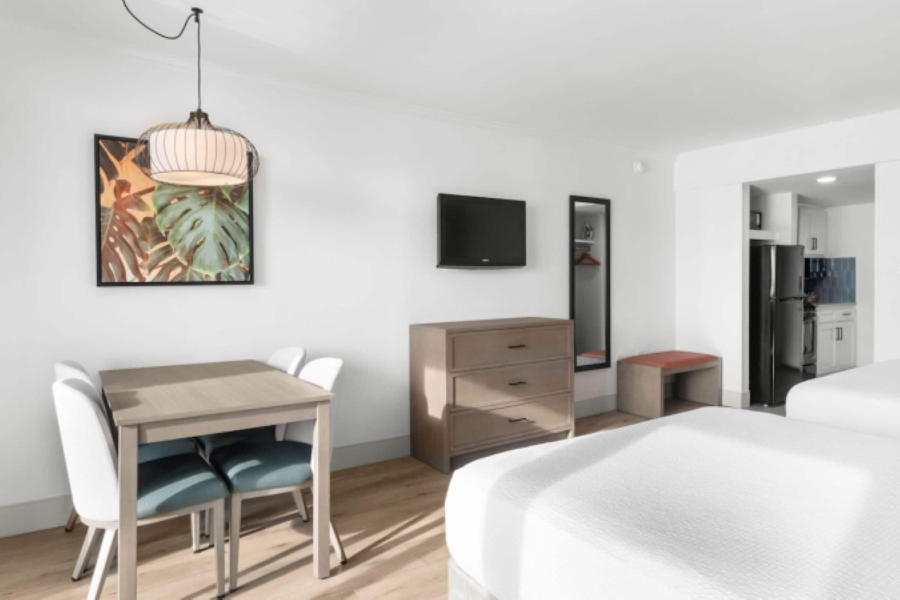 Una habitación de hotel moderna con cama, escritorio con sillas, cómoda, televisión y una cocina visible a través de una puerta abierta.