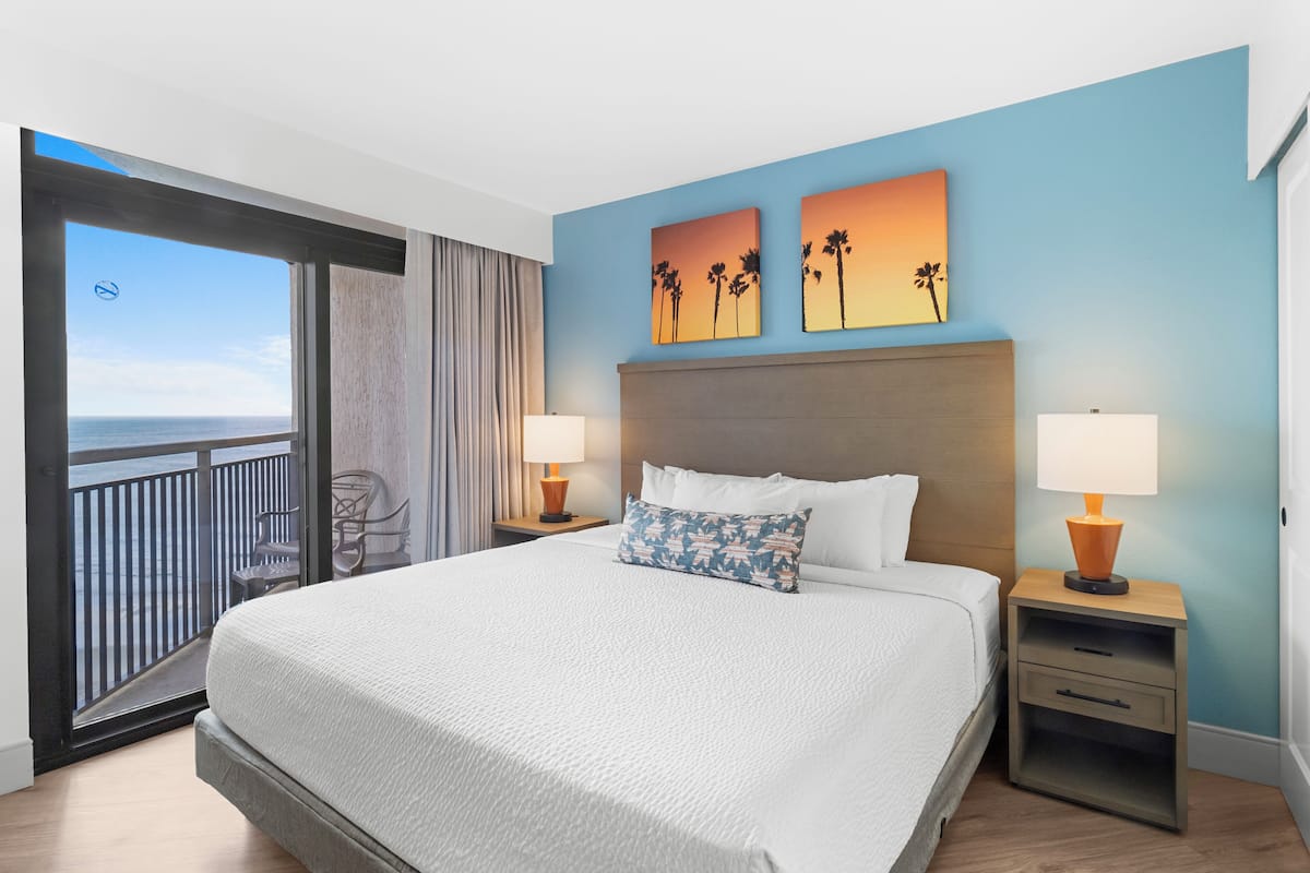 Quarto de hotel com uma cama grande, duas mesas de cabeceira, vista para o oceano e dois quadros de pôr do sol na parede.